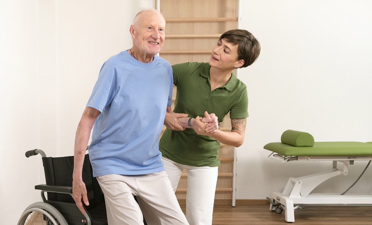 Therapeutin unterstützt älteren Mann beim Aufstehen aus dem Rollstuhl in einer ergotherapeutischen Praxis
